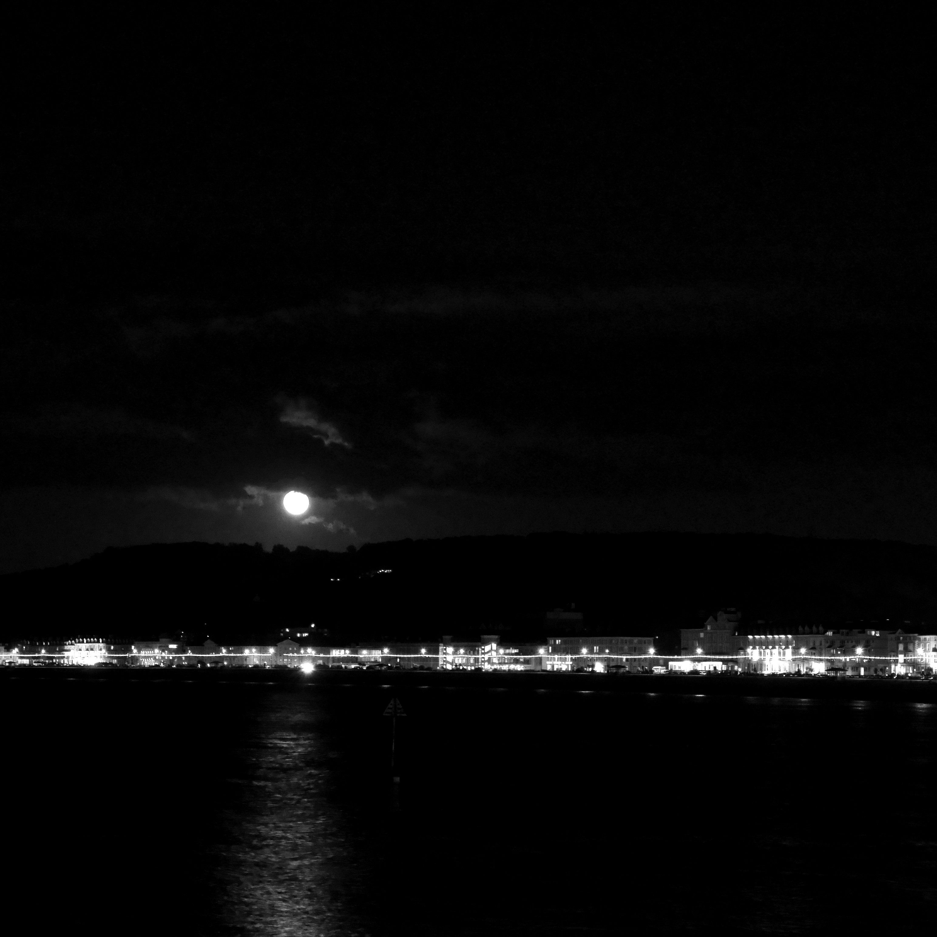 Very dark photo of a bay at night with a full moon shining over it, reflected in the water. Across the bay, street lights illimuninate a row of buildings.