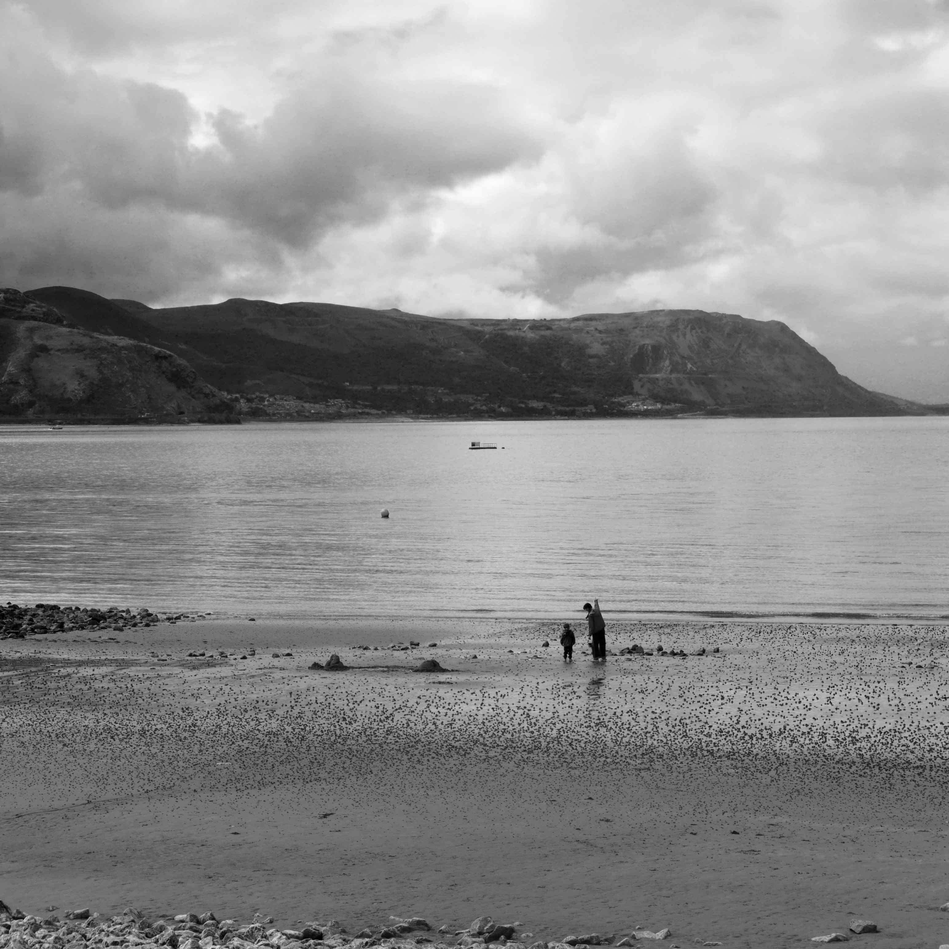 Wide shot of a boy and his mother alone on a sandy beach. Hills in background at the other side of the bay. The mother is pointing at something.