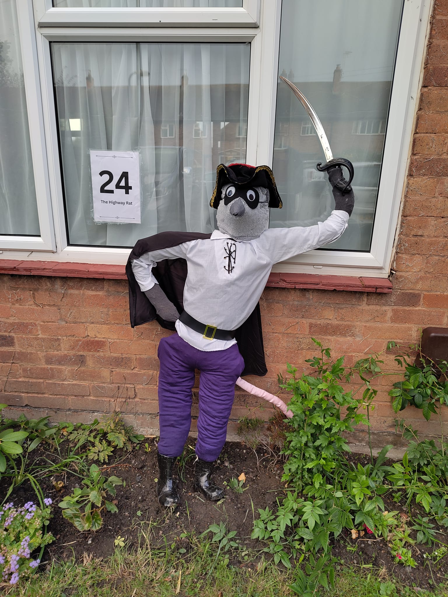 A Highway Rat scarecrow outside a houuse. He hoids a toy cutlass, and wears a white shirt, purple trousers, a mask, a cape, and a tricorn hat with a red feather