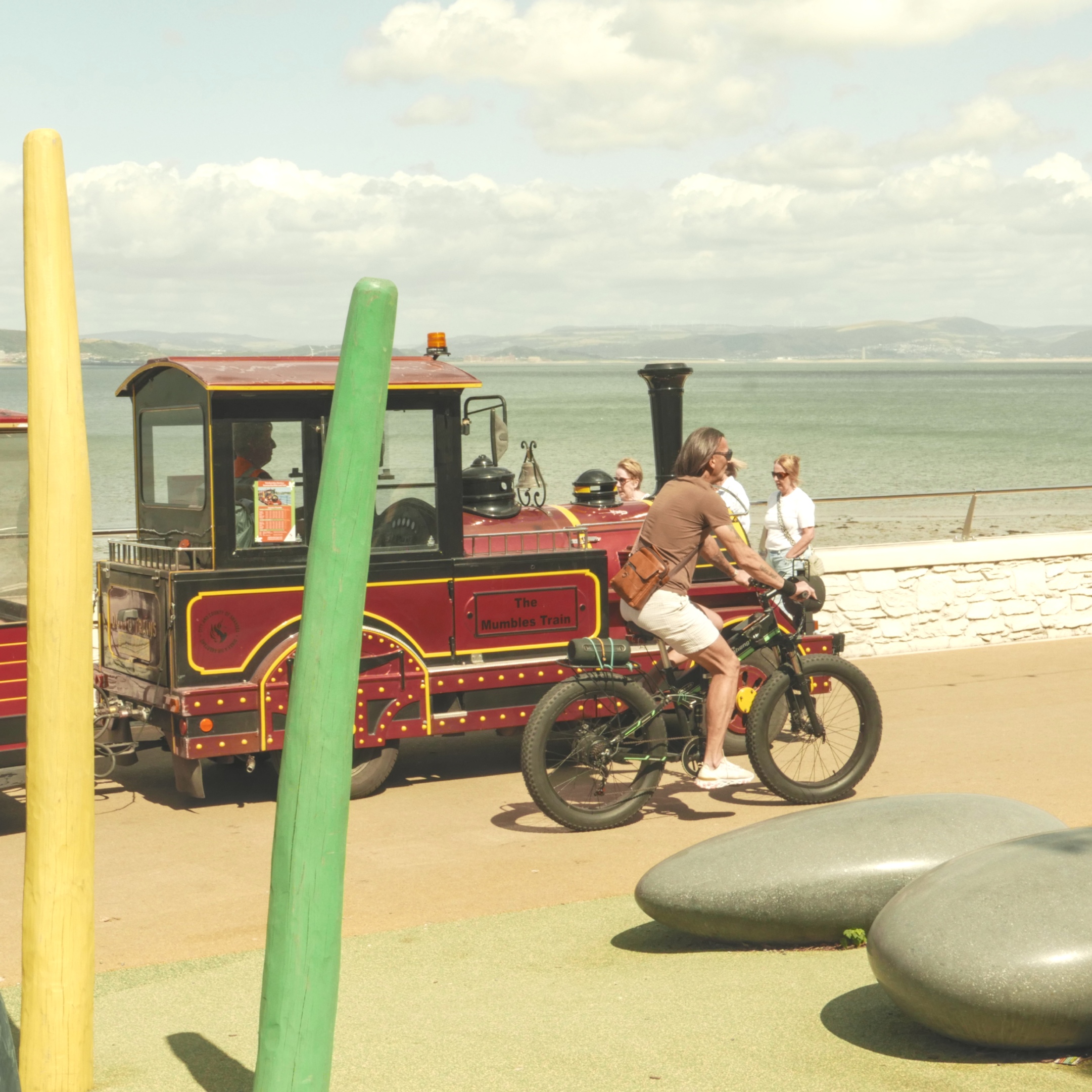 An image of the engine of a red land train on a promenade by the sea with a bicycle speeding by.