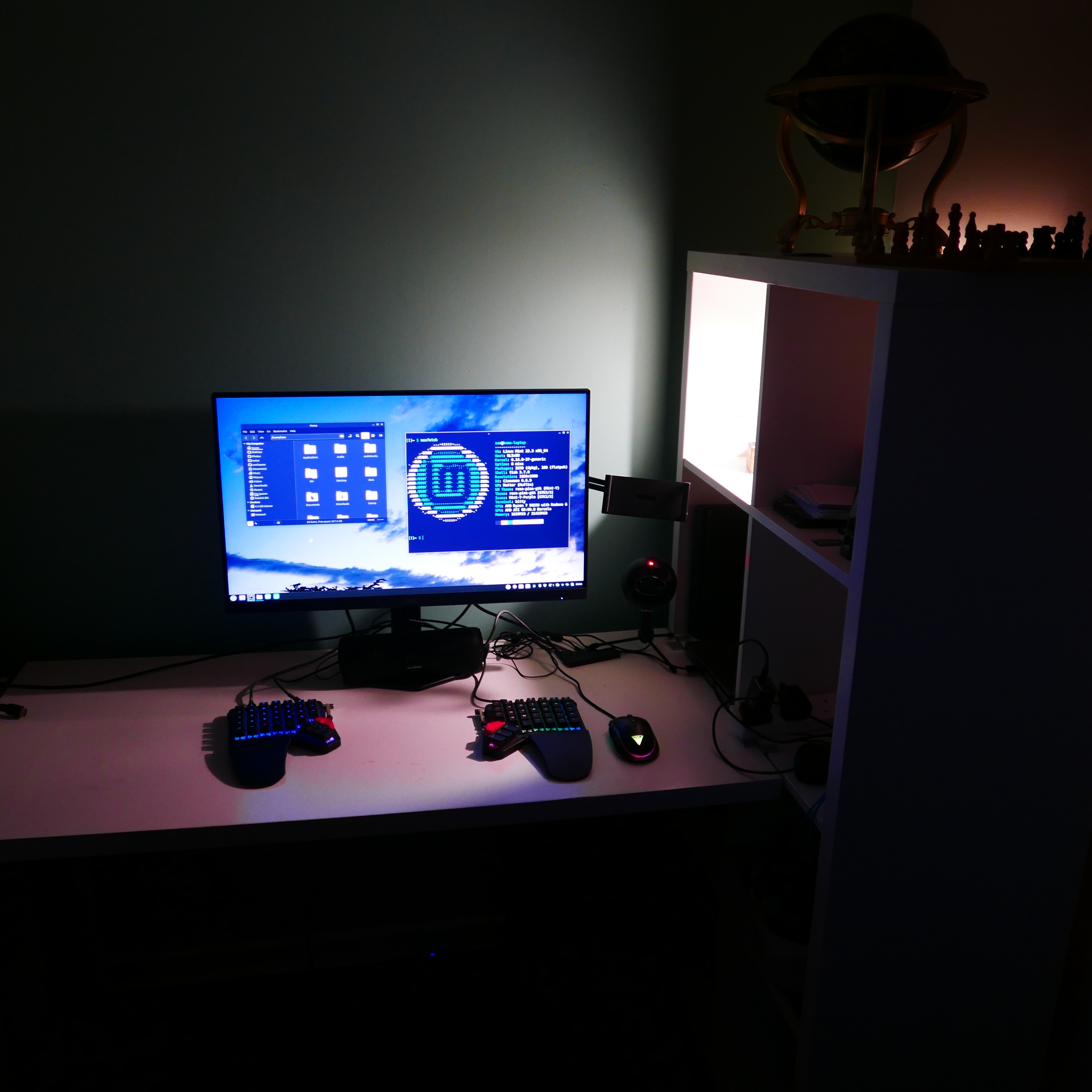 A white desk with a monitor, ZSA moonlander split ergonomic keyboard, mouse, speaker, and microphone visible. On a shelving unit by the desk is some kind of globe and a chess set.