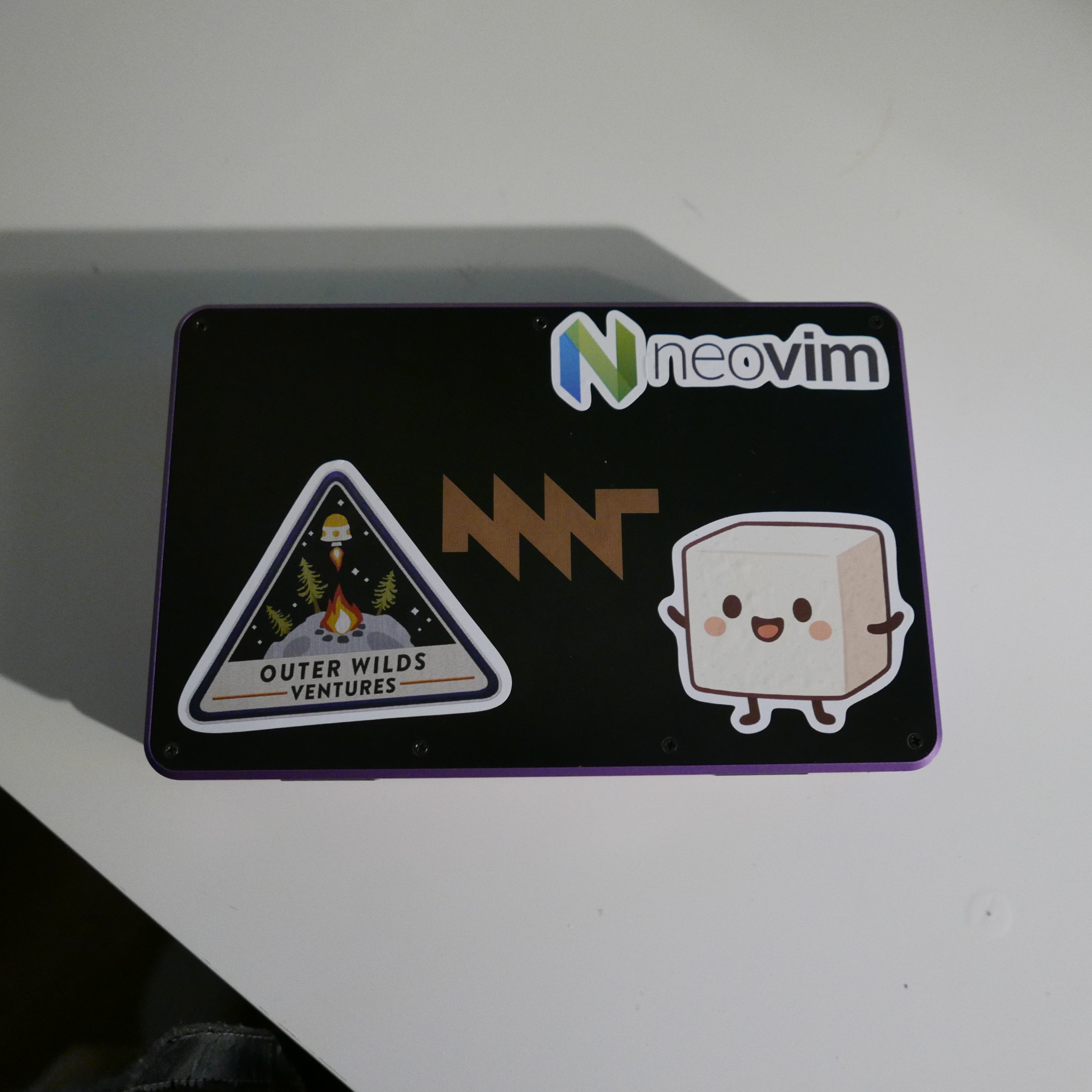 A close-up of the stickers on the laptop lid. One is a 'kawaii'-style tofu block, another is the Neovim logo, and finally, there is a sticker from the video game Outer Wilds. The MNT logo is also visible in the middle.